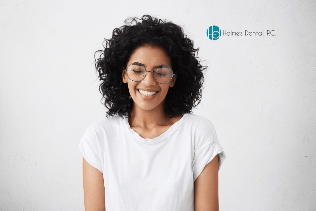 A smiling person with curly hair and glasses stands against a white background. They wear a white t-shirt, conveying a relaxed and happy mood. Holmes Dental, P.C. logo is in the top right.