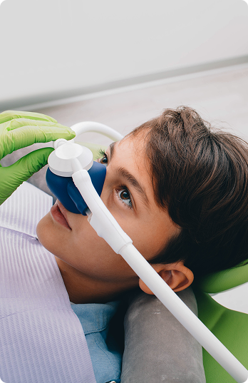 A child in a dental chair wears a blue nasal mask for sedation. A gloved hand adjusts the mask. The child appears calm, surrounded by clinical tools.