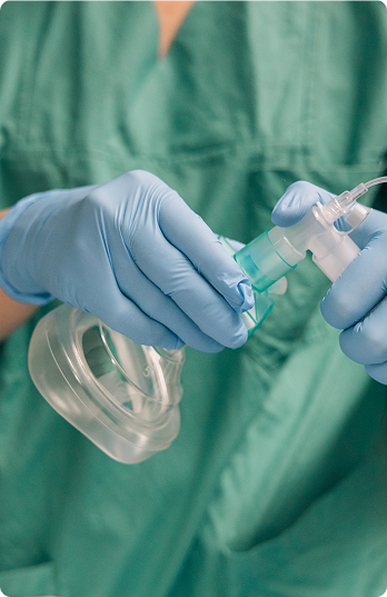 Hands wearing blue gloves hold a clear oxygen mask with green medical attire in the background, conveying a clinical and professional tone.