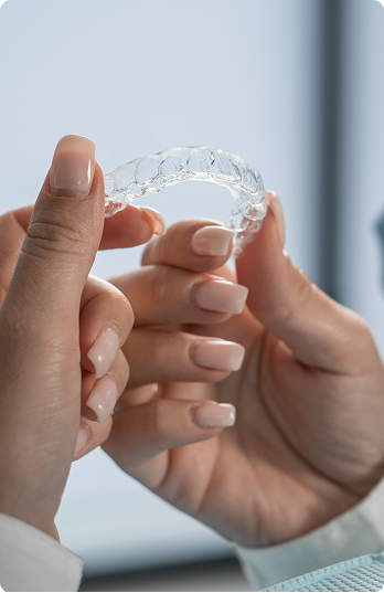 A person with manicured nails holds a clear dental aligner gently between their fingers. The background is softly blurred, suggesting a clinical setting.
