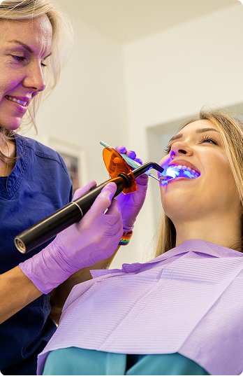 A dentist wearing purple gloves uses a curing light on a smiling patient in a dental chair. The setting is bright and professional, conveying care.