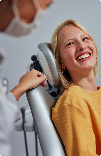 A woman in a yellow top smiles while sitting in a dentist's chair, looking at a healthcare professional who is leaning in, conveying a friendly, relaxed atmosphere.