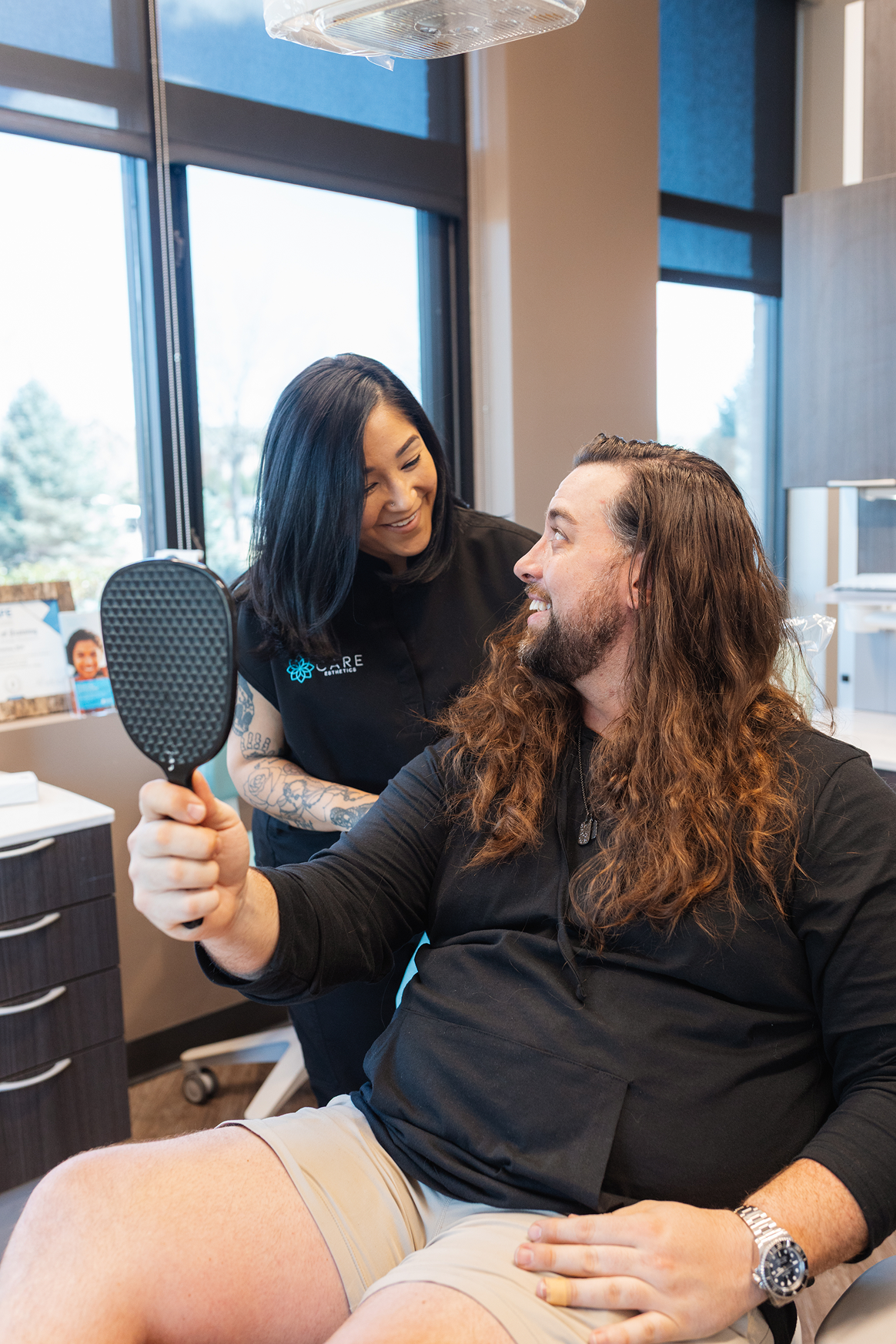 A woman with dark hair smiles at a man with long hair holding a mirror in a bright office. They seem pleased, creating a friendly and positive atmosphere.