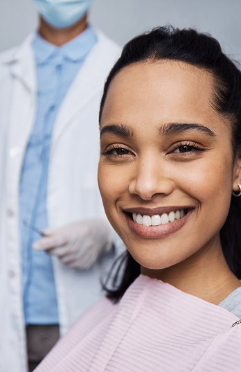 Smiling woman in a dental chair with a pink bib, and a dentist in the background wearing a mask and gloves, conveying a sense of care and professionalism.