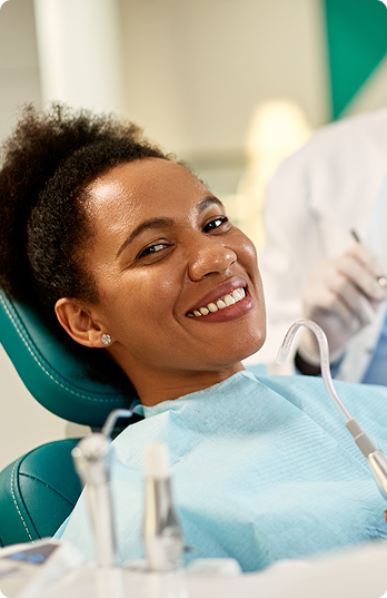 Smiling woman in a dental chair wearing a blue bib, with dental instruments nearby. A dentist in white coat appears blurred in the background.