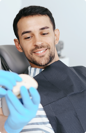 Smiling man in a dental chair, wearing a bib, looks at a set of teeth held by a person in blue gloves. The atmosphere is calm and reassuring.