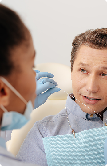 A dentist with gloves and a mask explains dental flossing to a patient in a clinic. The patient listens intently, with a dental bib, creating a calm atmosphere.