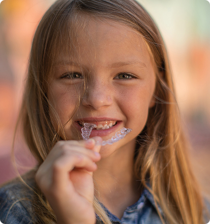 A smiling girl with long blond hair holds a clear dental retainer up to her mouth. She appears cheerful and confident against a blurred, warm-toned background.