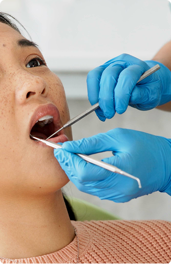 A patient in an orange sweater receives a dental checkup. A dentist in blue gloves uses a mirror and probe to examine her open mouth.