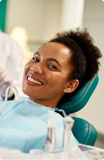 A woman smiling confidently while seated in a dental chair, wearing a blue bib. The setting is a dental clinic, conveying a sense of comfort and trust.