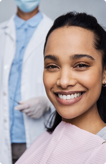 Smiling woman in a dental chair with a pink bib, dentist in the background wearing a mask and gloves. The atmosphere is calm and professional.