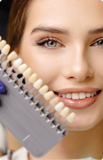 A smiling woman in a dental office holds a tooth color palette near her teeth. A gloved hand holds the palette, suggesting a tooth whitening consultation.